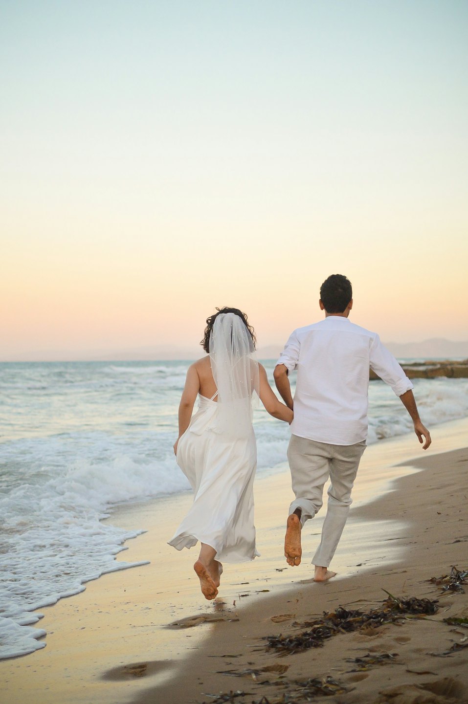 Recién casados corren descalzos por la orilla de la playa de arena, tomados de la mano.
